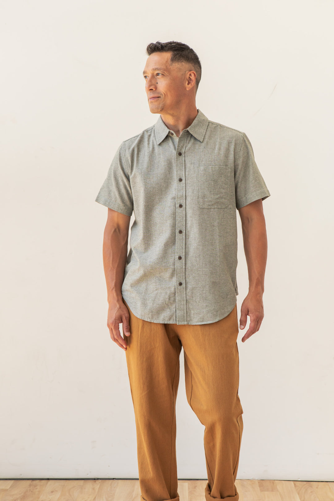 A man wears the Bridge & Burn Malcolm Shirt in Sage Chambray and tan pants, standing on a light wood floor with an off-white background, looking to his left with a calm expression.