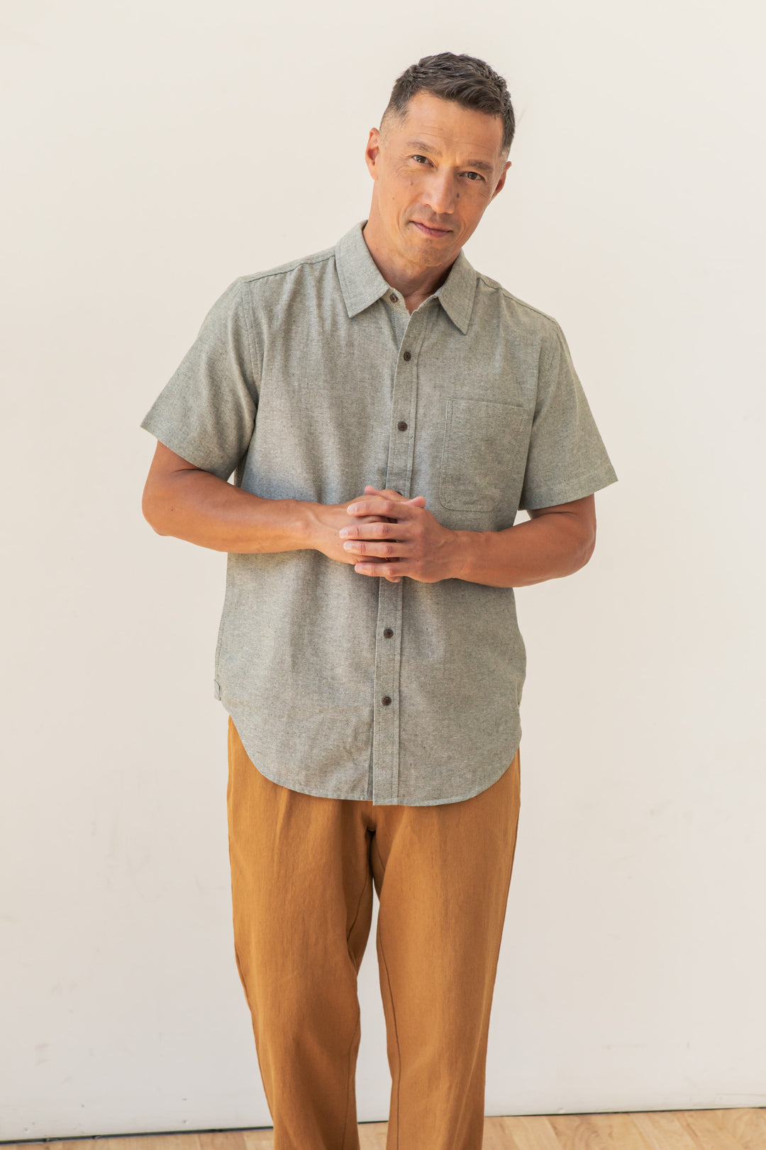 A man with short dark hair stands against a light background, wearing the Bridge & Burn Malcolm Shirt in Sage Chambray and mustard pants. He clasps his hands in front of him and looks at the camera.