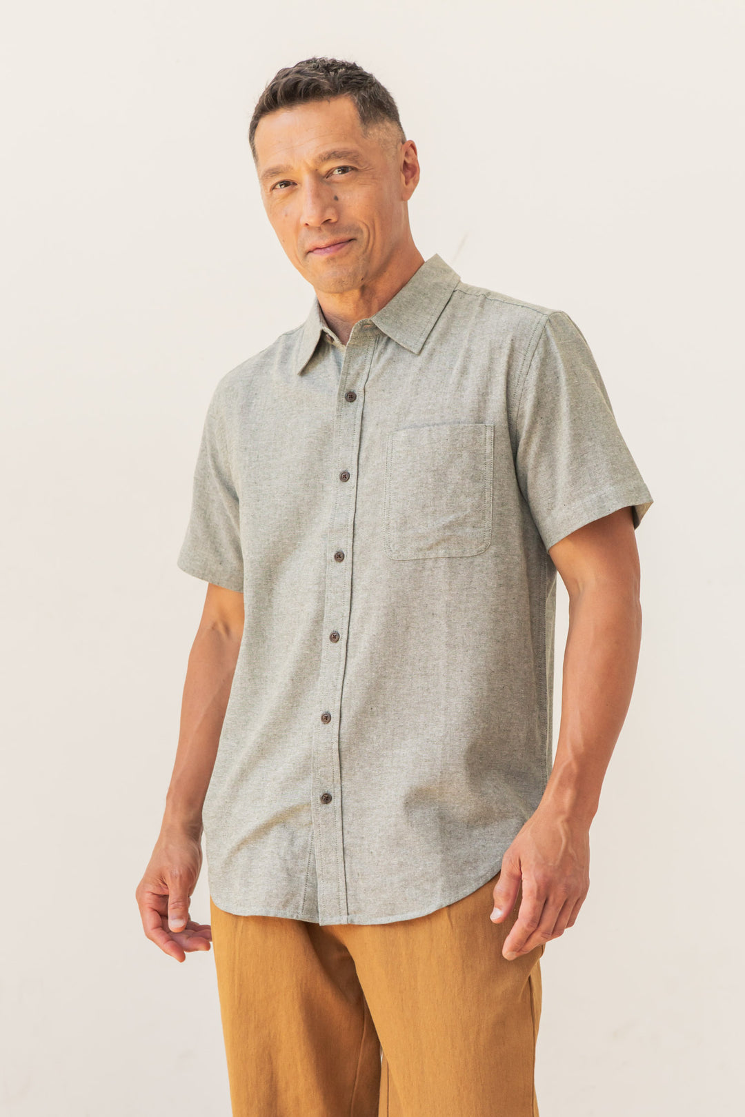 A man with short dark hair and a neutral expression stands against a plain background, wearing the Bridge & Burn Malcolm Shirt in Sage Chambray— a sustainable men's short-sleeve button-up with front pocket—paired with tan pants.