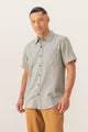 A man with short dark hair and a neutral expression stands against a plain background, wearing the Bridge & Burn Malcolm Shirt in Sage Chambray— a sustainable men's short-sleeve button-up with front pocket—paired with tan pants.