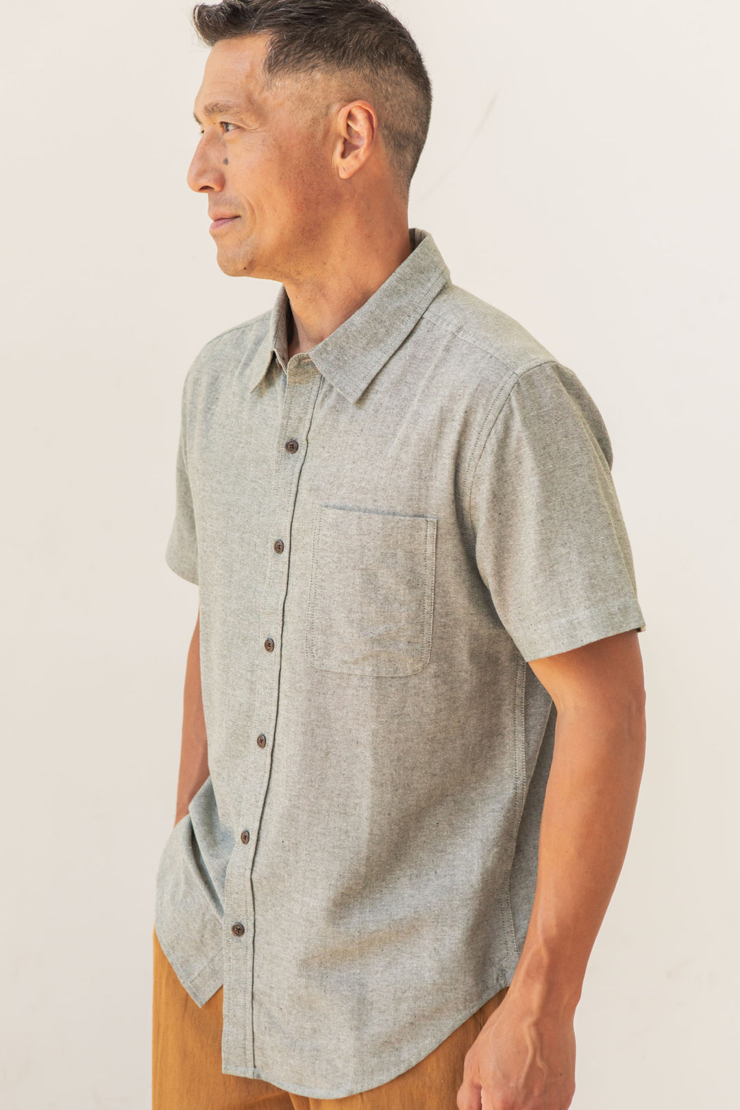 A man wears the Bridge & Burn Malcolm Shirt in Sage Chambray, a light gray recycled cotton short sleeve button-up with a chest pocket, standing against an off-white background as he smiles and looks to his left.