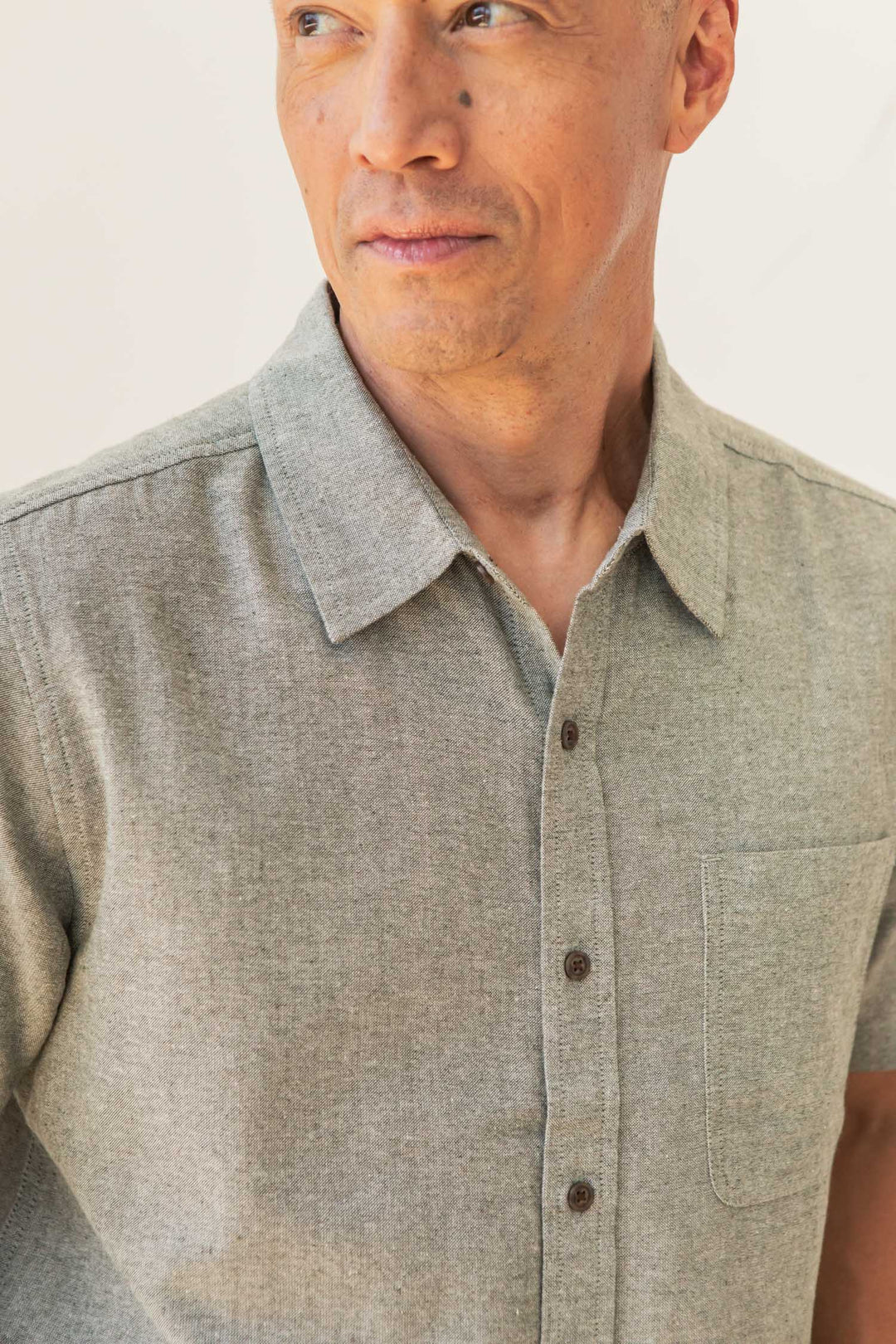 A man wearing the Bridge & Burn Malcolm Shirt in Sage Chambray—a light gray recycled cotton short sleeve button-up with a collar and chest pocket—looks to the side against a neutral background.