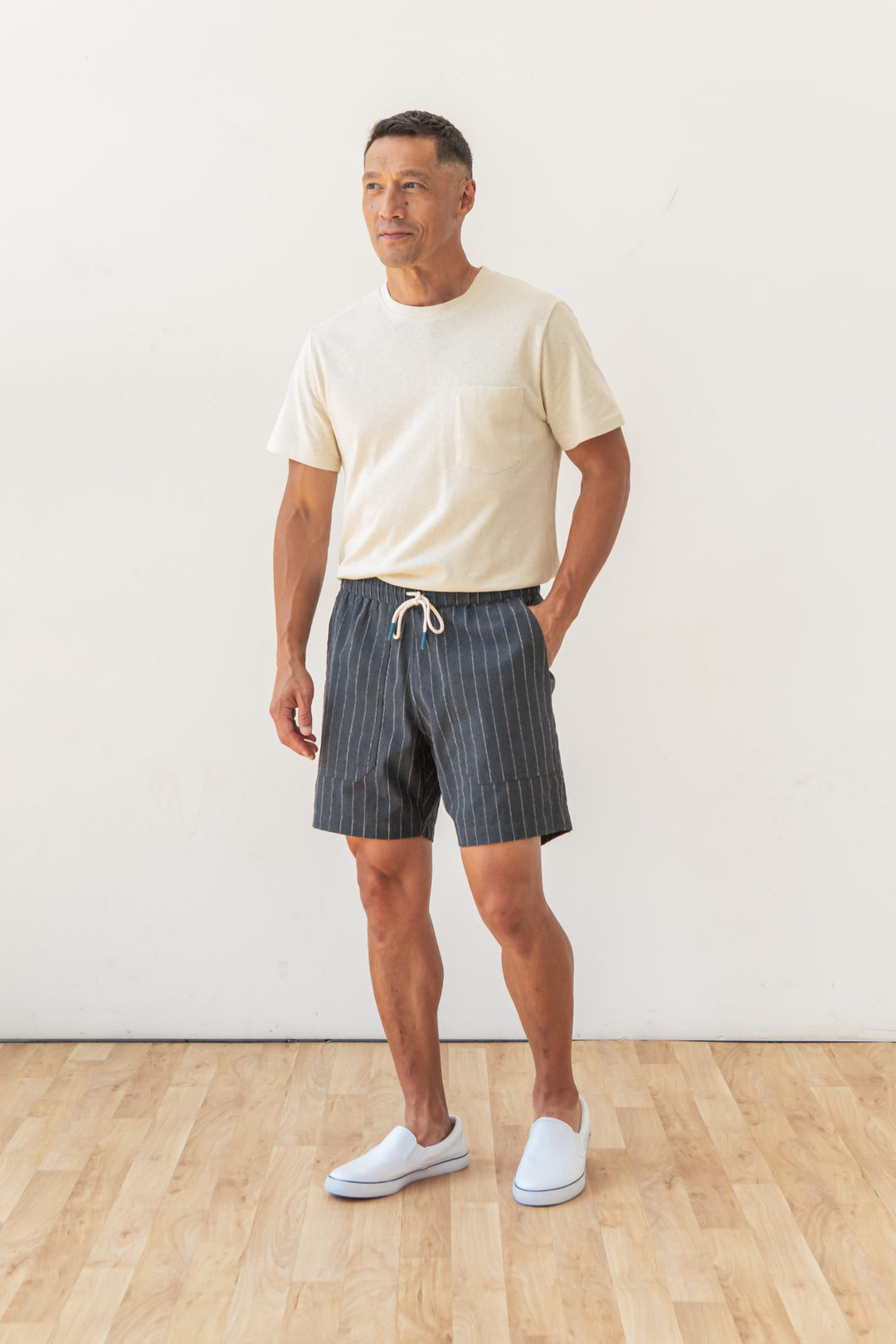 A man stands indoors on a wooden floor against a plain white background, wearing Bridge & Burn's Noah Short in Charcoal Stripe, a light beige t-shirt, and white slip-on shoes, with his left hand in his pocket.
