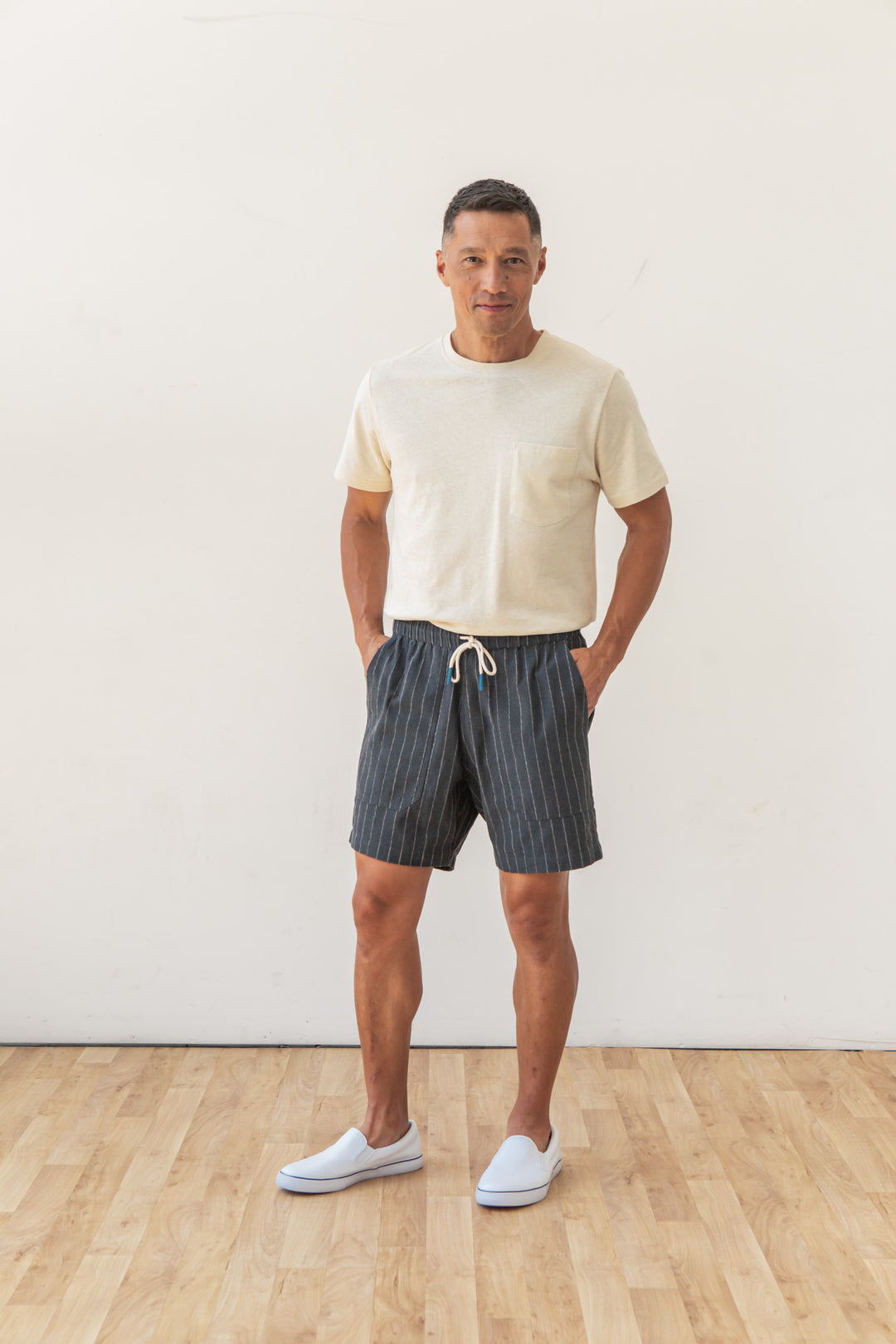 A man stands indoors on a wooden floor, wearing Bridge & Burn's Noah Short in Charcoal Stripe, paired with a cream t-shirt and white slip-on shoes. He faces forward with hands in pockets against a plain white wall.