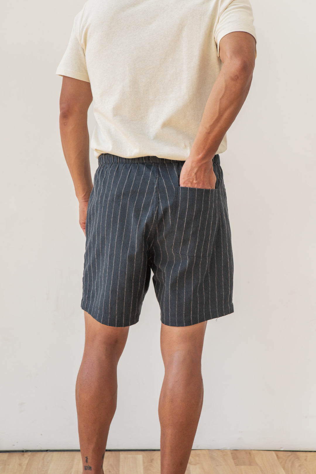 A person stands indoors facing away, wearing a cream short-sleeve shirt and Bridge & Burn’s Noah Short in Charcoal Stripe—dark pinstriped cotton-linen shorts with an elastic waist. Hands are tucked in pockets; the background is plain and light-colored.