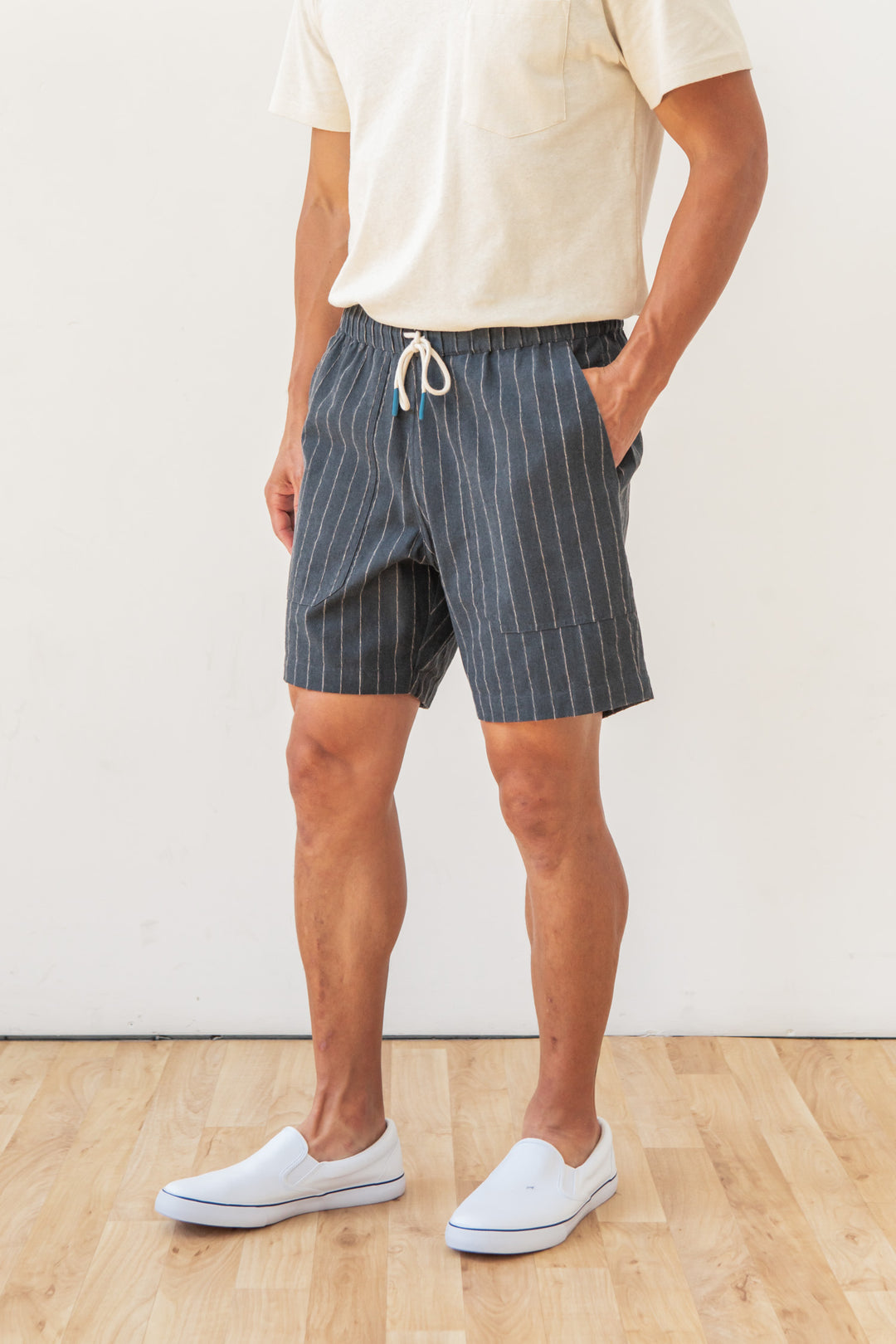 A person wearing a cream t-shirt, Bridge & Burn Noah Short in Charcoal Stripe, and white slip-on shoes stands on a light wooden floor against a plain white wall.
