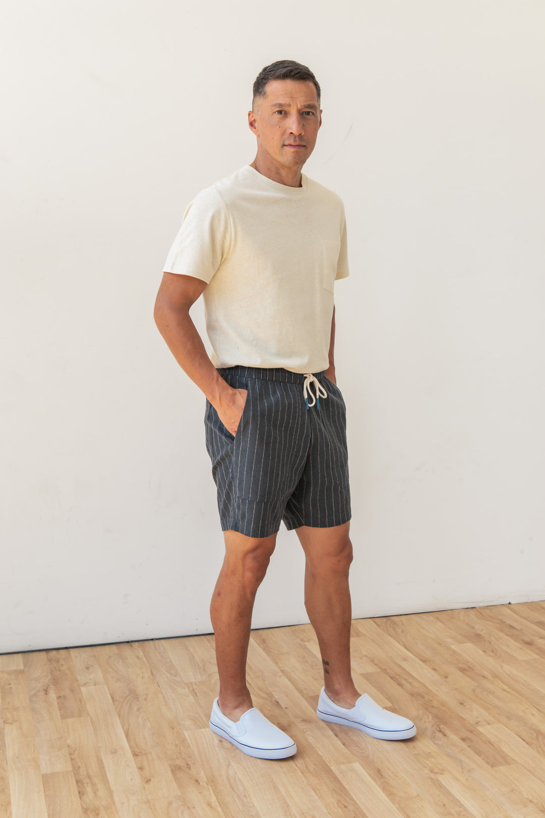 A man stands indoors on a wooden floor against a white wall, wearing the Bridge & Burn Noah Short in Charcoal Stripe with an elastic waist and drawstring, a light beige T-shirt, and white slip-on shoes. His hands are in his pockets.