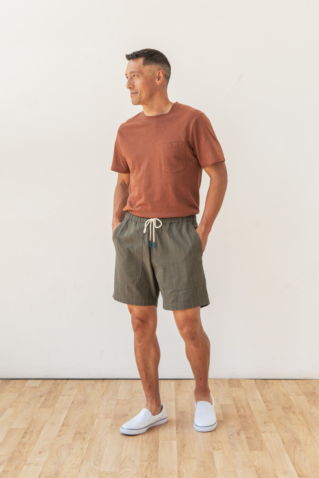 A man stands on a wooden floor wearing Bridge & Burn’s Noah Short in Olive Stripe—relaxed, pull-on cotton-linen blend shorts with an elastic waist. He has his hands in his pockets, white slip-on shoes, and smiles while looking left.