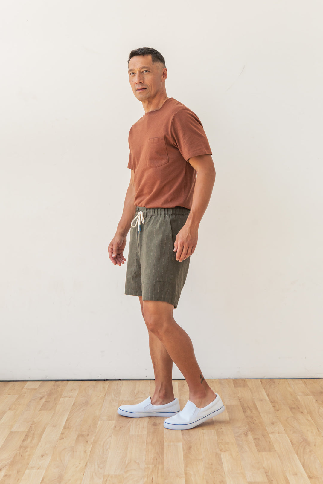 A man stands indoors on a wooden floor against a plain white wall, wearing Bridge & Burn's Noah Short in Olive Stripe with an elastic waist, a rust t-shirt, and white slip-on shoes. He faces slightly to the side and looks at the camera.