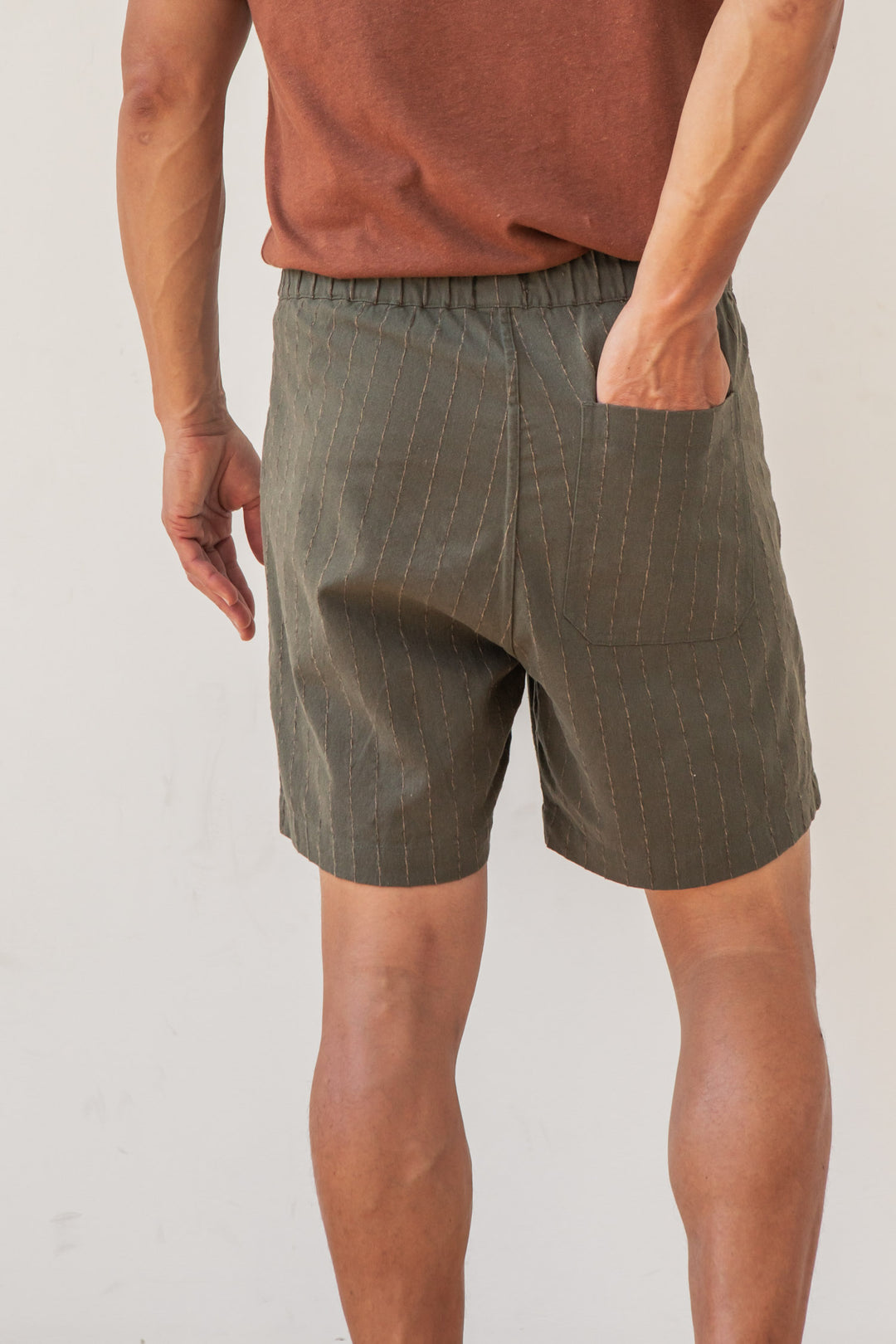 A person in Bridge & Burn’s Noah Short in Olive Stripe stands facing away with one hand in their back pocket, set against a plain white background.