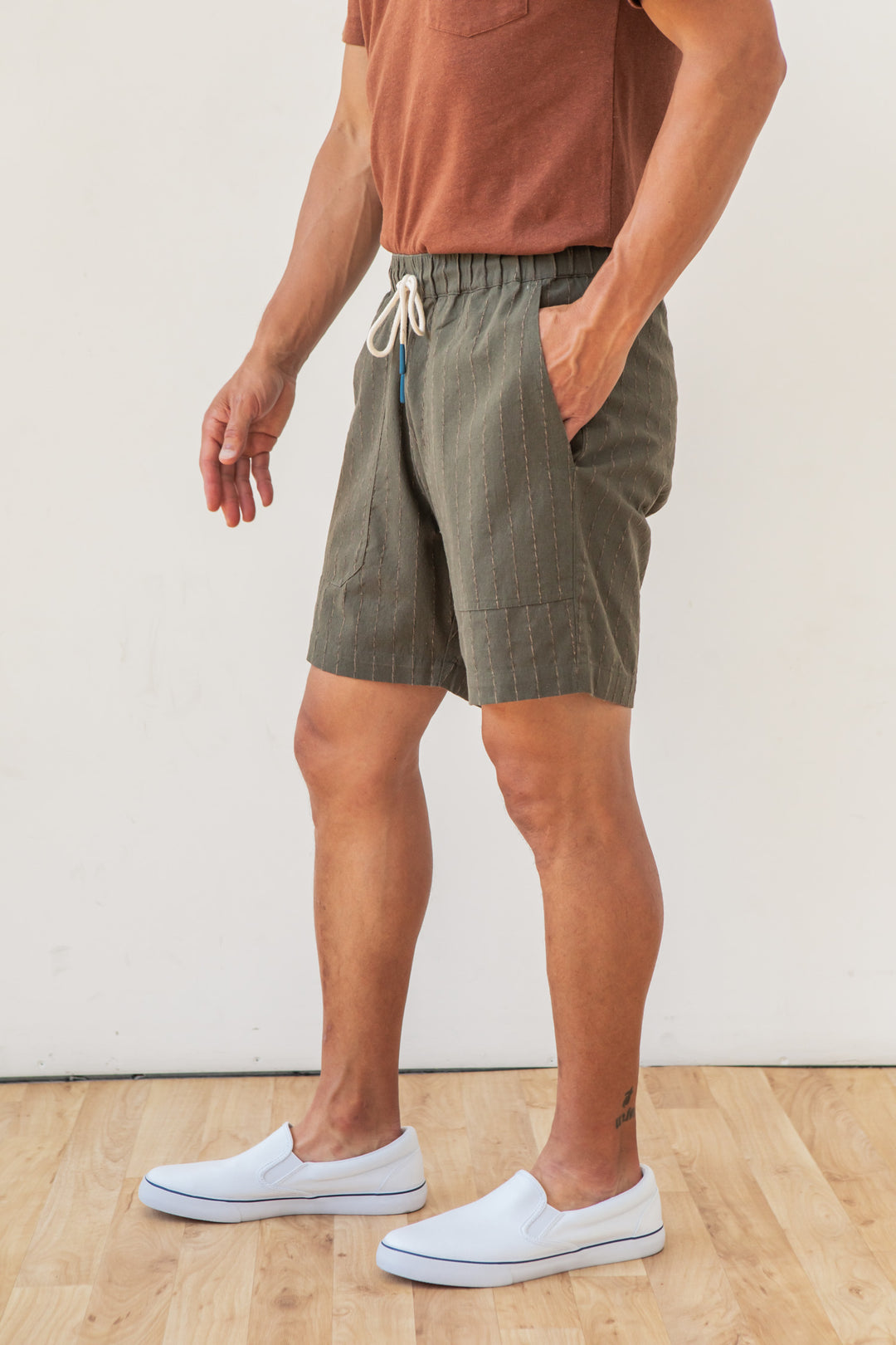 A person wearing a rust-colored t-shirt, white slip-on shoes, and Bridge & Burn's Noah Short in Olive Stripe stands on a light wood floor against an off-white wall.