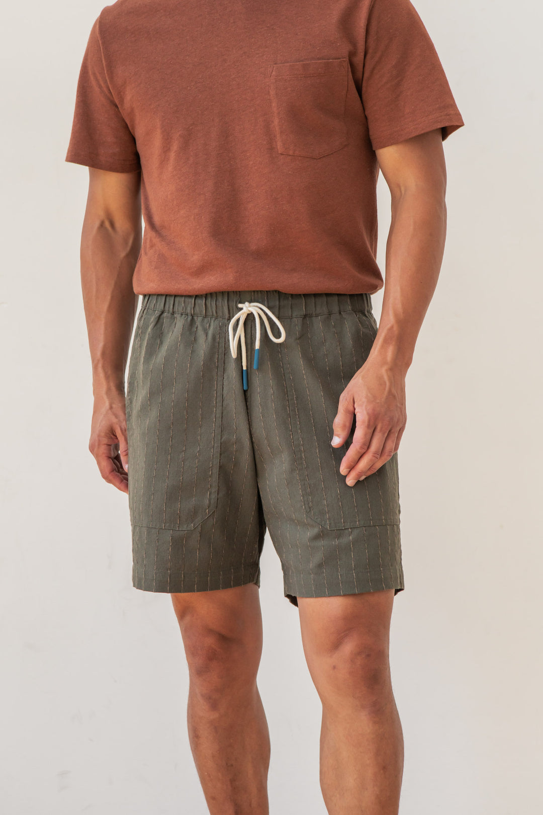 A person wearing the Bridge & Burn Noah Short in Olive Stripe, featuring subtle vertical stitching, stands against a plain light background. Their face is not visible.