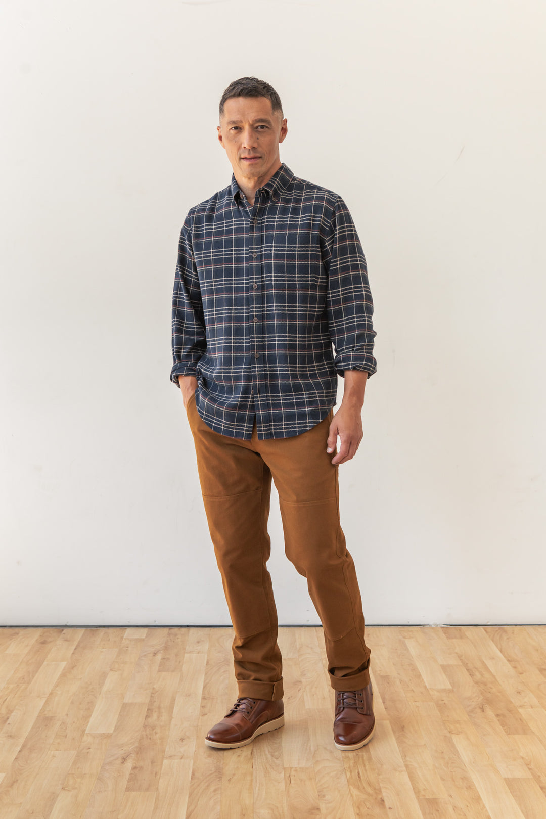 A man stands indoors on a wooden floor against a white wall, wearing the Bridge & Burn Sutton Slim Shirt in Dusk Herringbone Plaid, paired with brown pants and shoes, with his left hand in his pocket and a neutral expression.