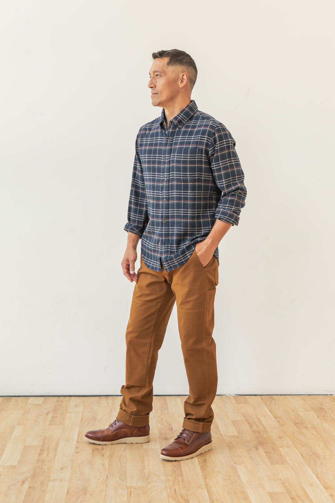 A man stands on a wooden floor against a white wall, wearing the Bridge & Burn Sutton Slim Shirt in Dusk Herringbone Plaid, with brown pants and leather shoes, hands in pockets, looking to the side.