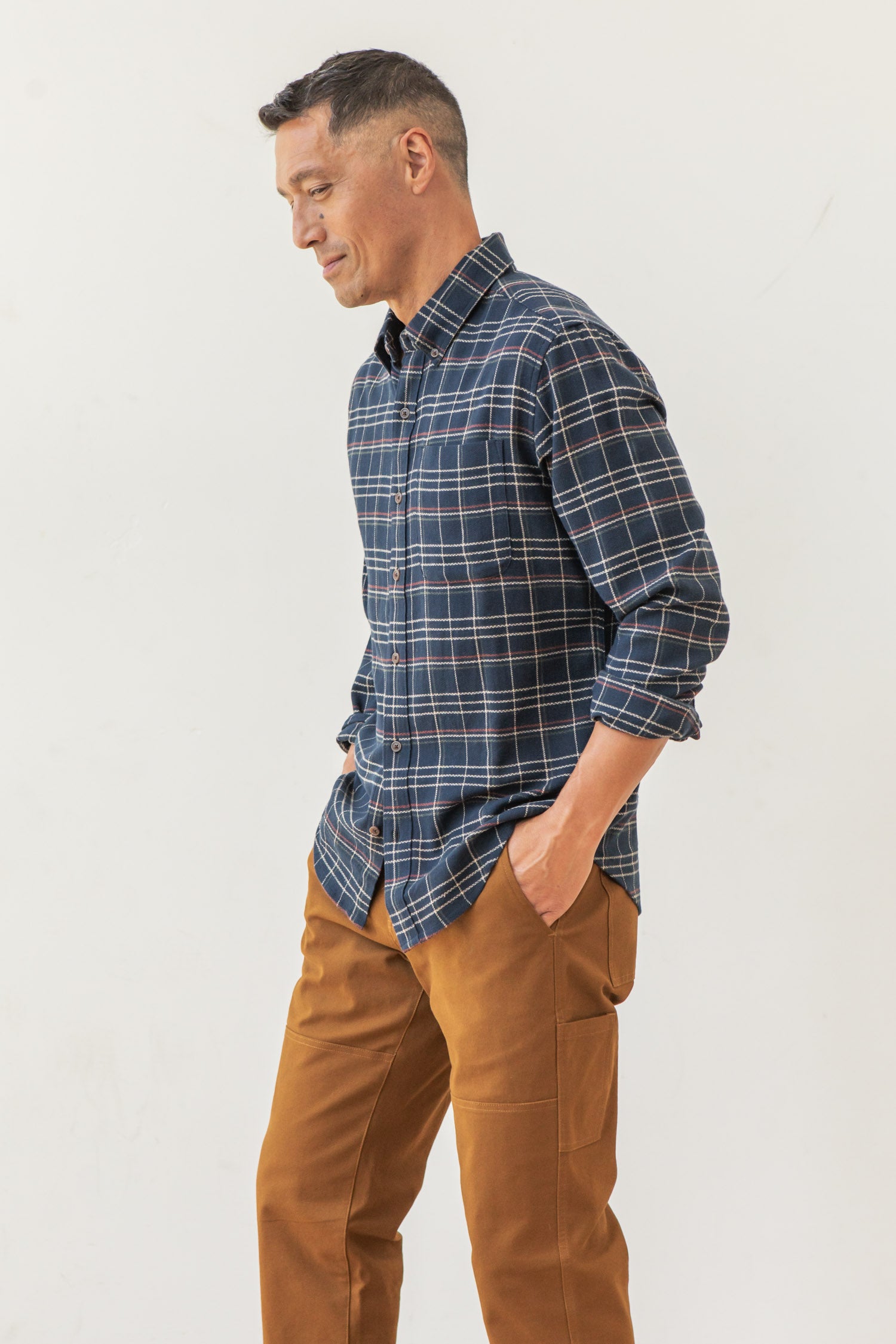 A man stands sideways against a white background, hands in pockets and relaxed, wearing the Bridge & Burn Sutton Slim Shirt in Dusk Herringbone Plaid with brown pants.