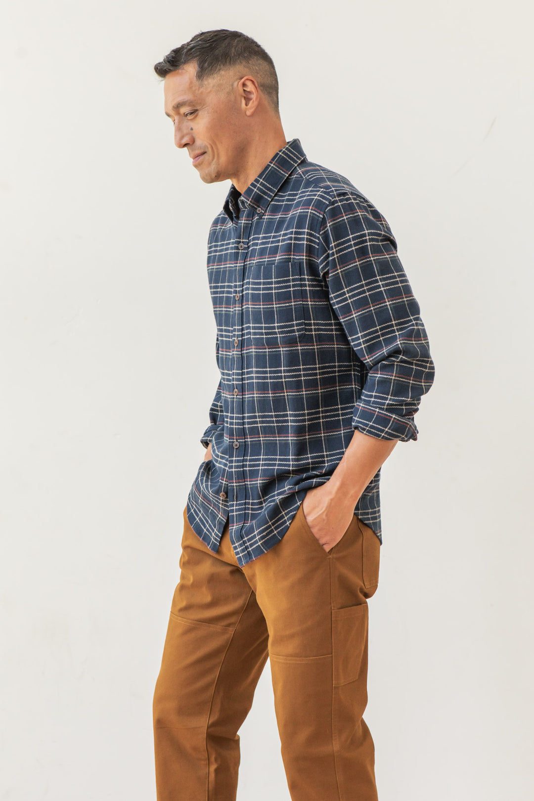 A man stands sideways against a white background, hands in pockets and relaxed, wearing the Bridge & Burn Sutton Slim Shirt in Dusk Herringbone Plaid with brown pants.