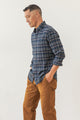 A man stands sideways against a white background, hands in pockets and relaxed, wearing the Bridge & Burn Sutton Slim Shirt in Dusk Herringbone Plaid with brown pants.