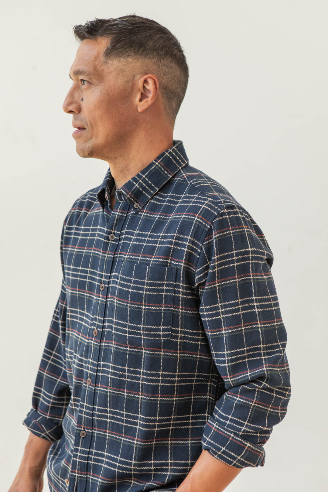 A man with short dark hair, wearing the Bridge & Burn Sutton Slim Shirt in Dusk Herringbone Plaid, stands facing left against a plain light background.