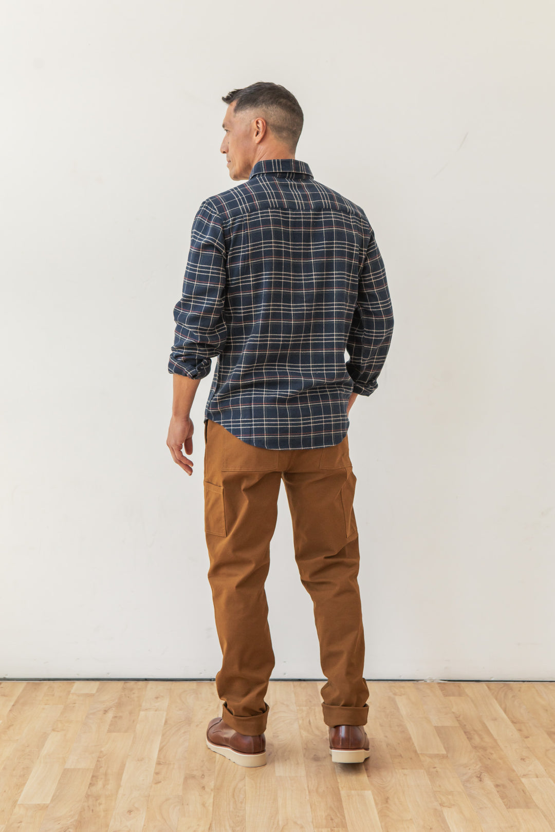 A man stands on a wooden floor facing away, wearing the Bridge & Burn Sutton Slim Shirt in Dusk Herringbone Plaid, along with brown pants and shoes. The backdrop is a plain white wall, creating a clean, minimalist look.