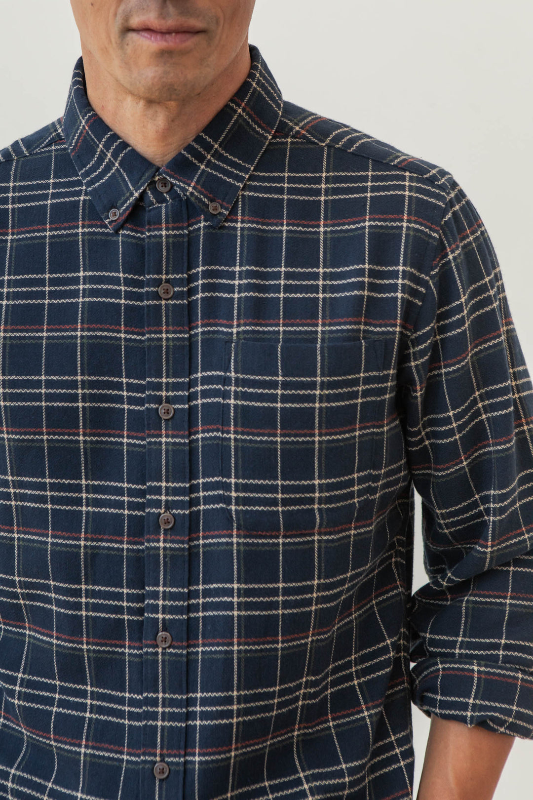 A person wears the Bridge & Burn Sutton Slim Shirt in Dusk Herringbone Plaid, a navy slim-fit button-up with red, white, and beige lines. The image shows their upper body from chest to just above the mouth, with one sleeve slightly rolled up.