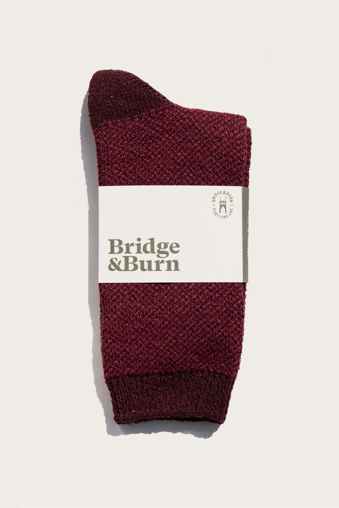 A single Helix Sock in burgundy from Bridge & Burn is folded and wrapped with a white brand label, set against a plain light background.