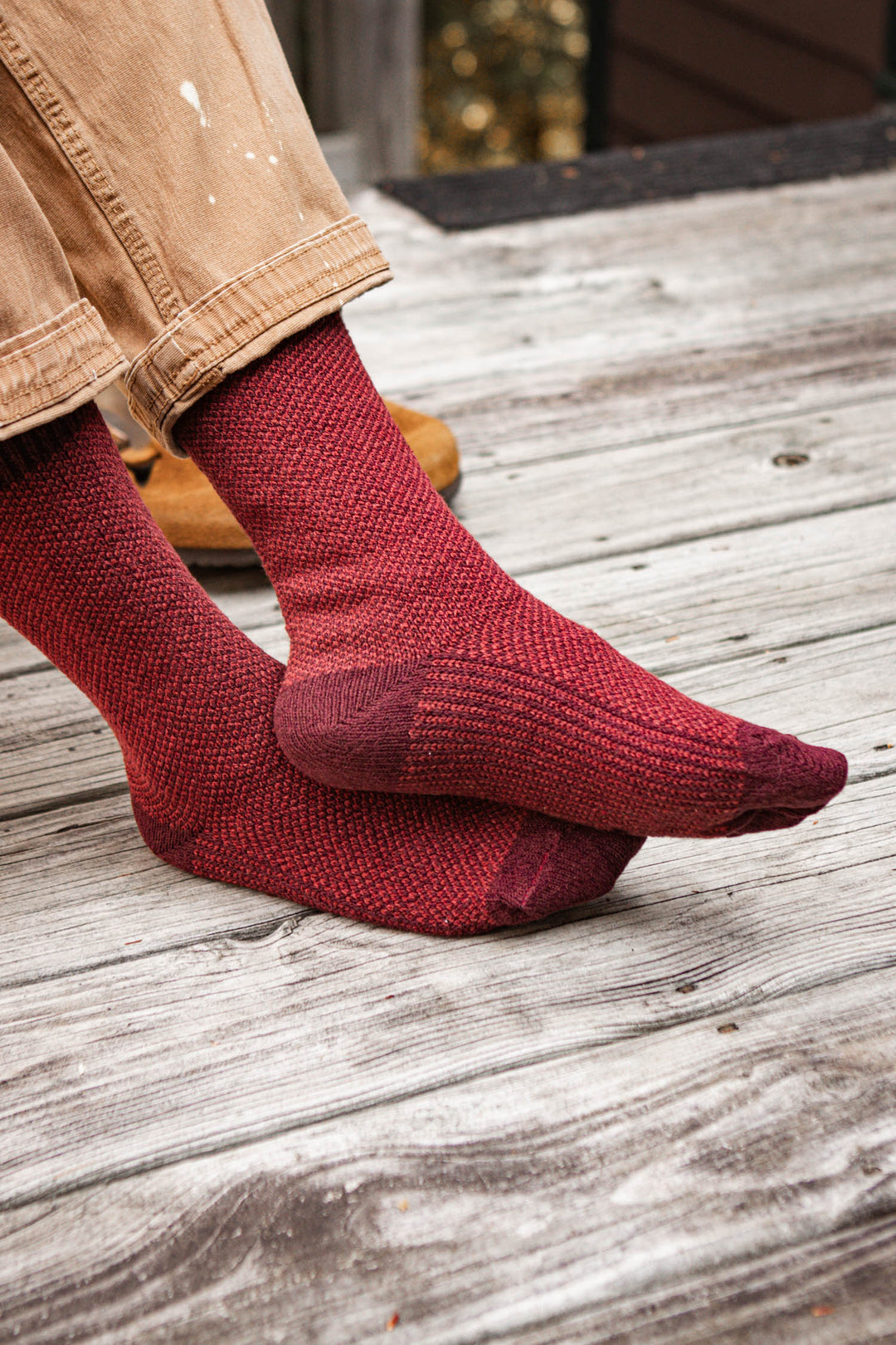 A person wearing tan pants and Bridge & Burn Helix Socks in Burgundy sits cross-legged on a weathered wooden deck.