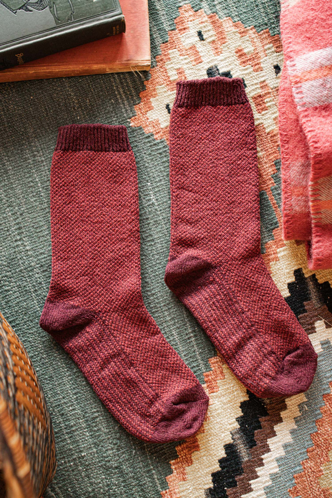 A pair of Bridge & Burn Helix Socks in burgundy lies side by side on a patterned rug, with parts of a book and a pink blanket nearby.
