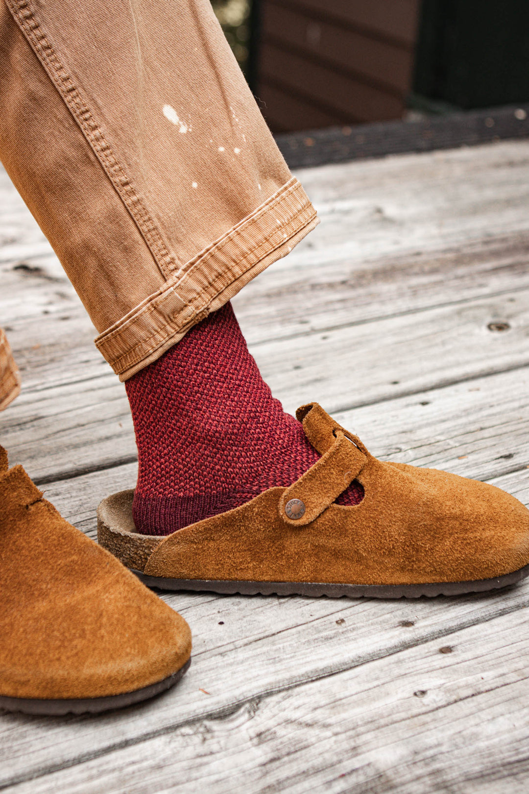 A person wearing light brown pants, Bridge & Burn Helix Sock in Burgundy, and tan suede clogs stands on a weathered wooden deck.