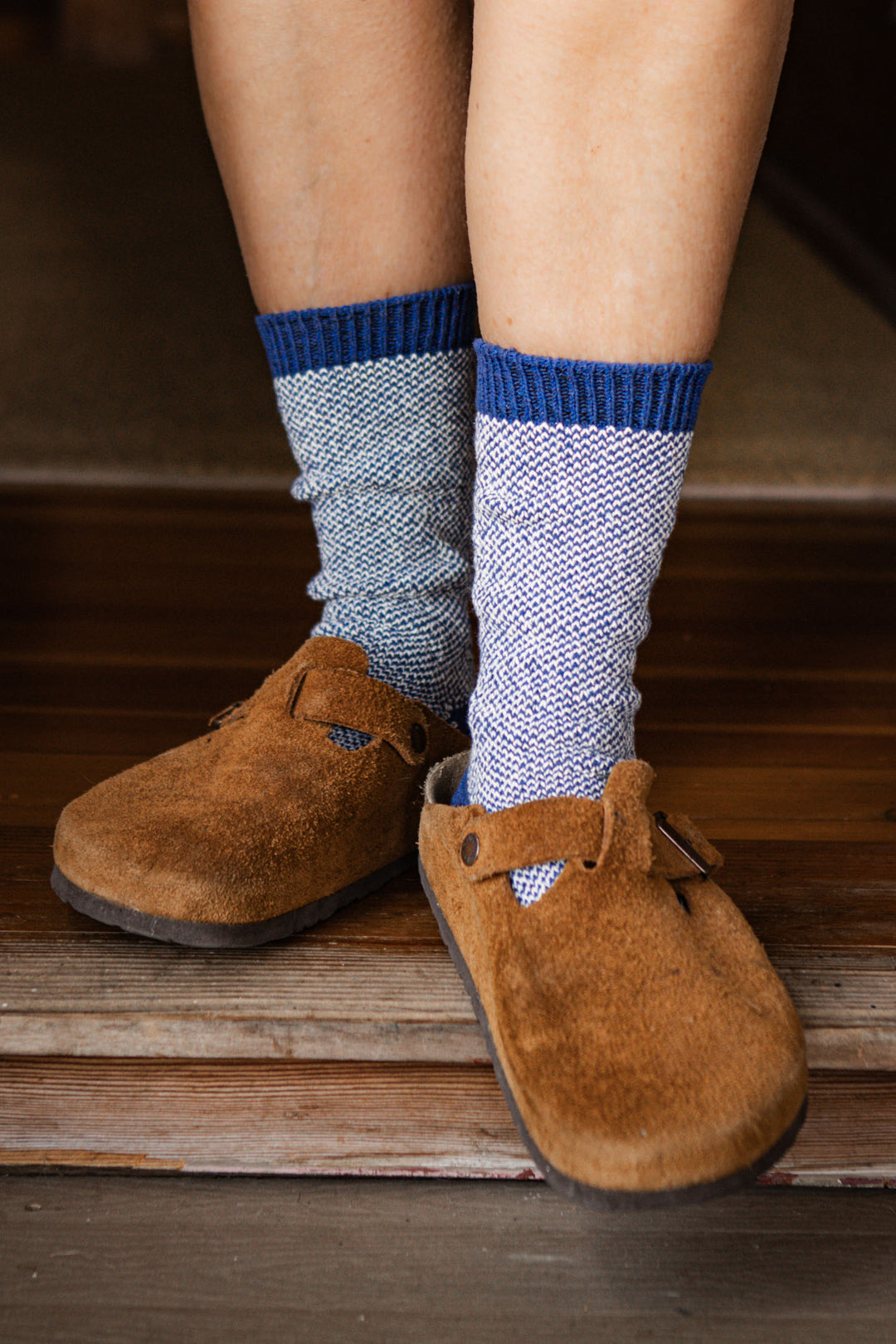 A person wearing Bridge & Burn's Helix Sock in Navy stands on a wooden floor near a step, paired with brown suede slip-on clogs.