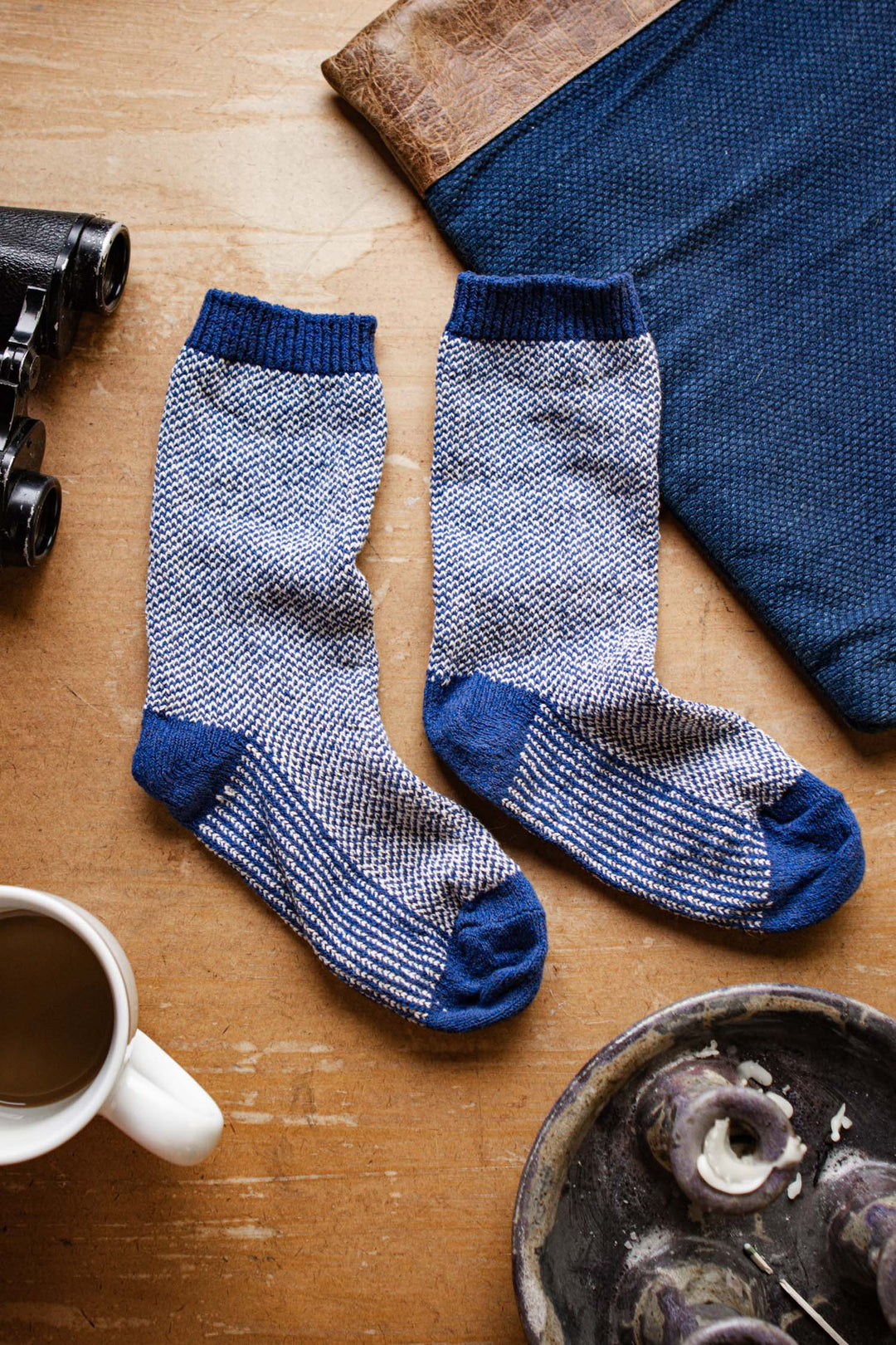 A pair of Bridge & Burn Helix Socks in navy rests on a wooden surface beside a cup of coffee, binoculars, and a blue cloth with a leather corner.
