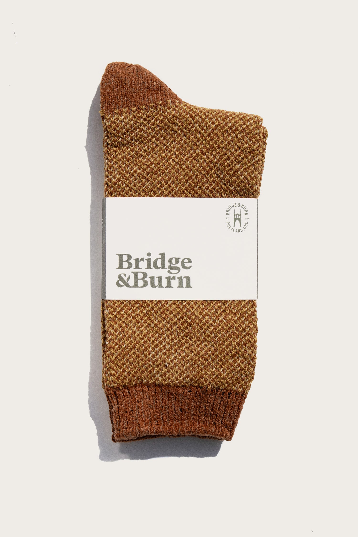 A pair of Helix Sock / Ochre by Bridge & Burn, textured brown and neatly folded with a white label featuring the brand name in bold letters, displayed against a plain white background.
