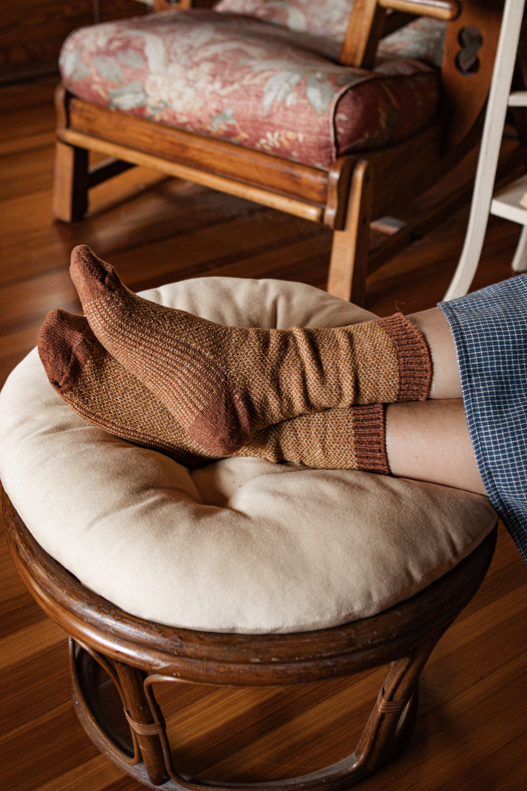 Someone wearing Bridge & Burn’s Helix Sock in Ochre relaxes with their feet on a round, cushioned footstool amid warm wooden furniture and flooring in a cozy, welcoming space.