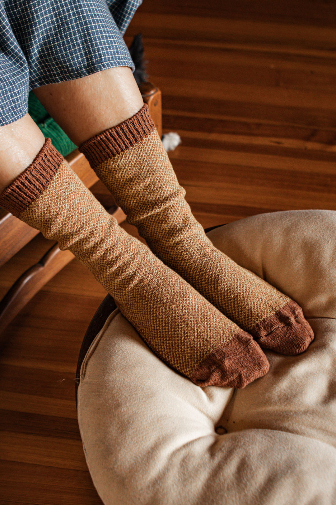 Someone wearing Bridge & Burn's Helix Sock in Ochre, made in the USA, rests their feet on a plush round beige cushion atop a wooden floor.