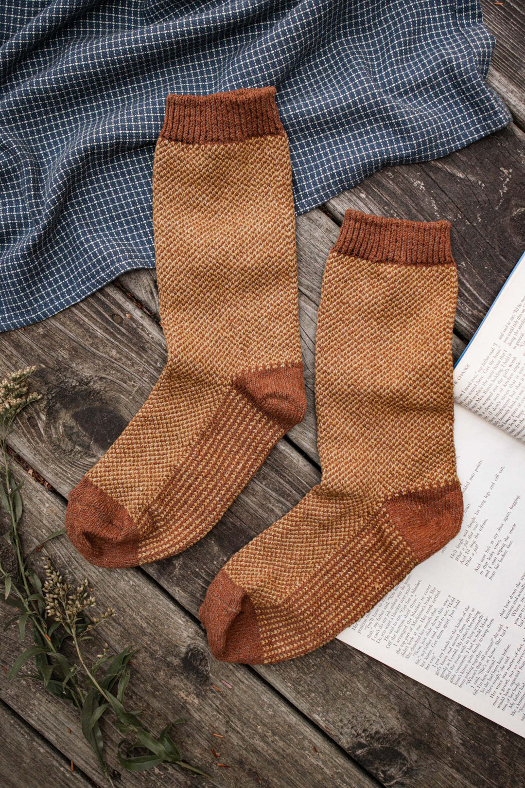 A pair of Bridge & Burn Helix Sock in Ochre rests on a rustic wooden surface next to an open book, dried sprigs, and blue checkered fabric.