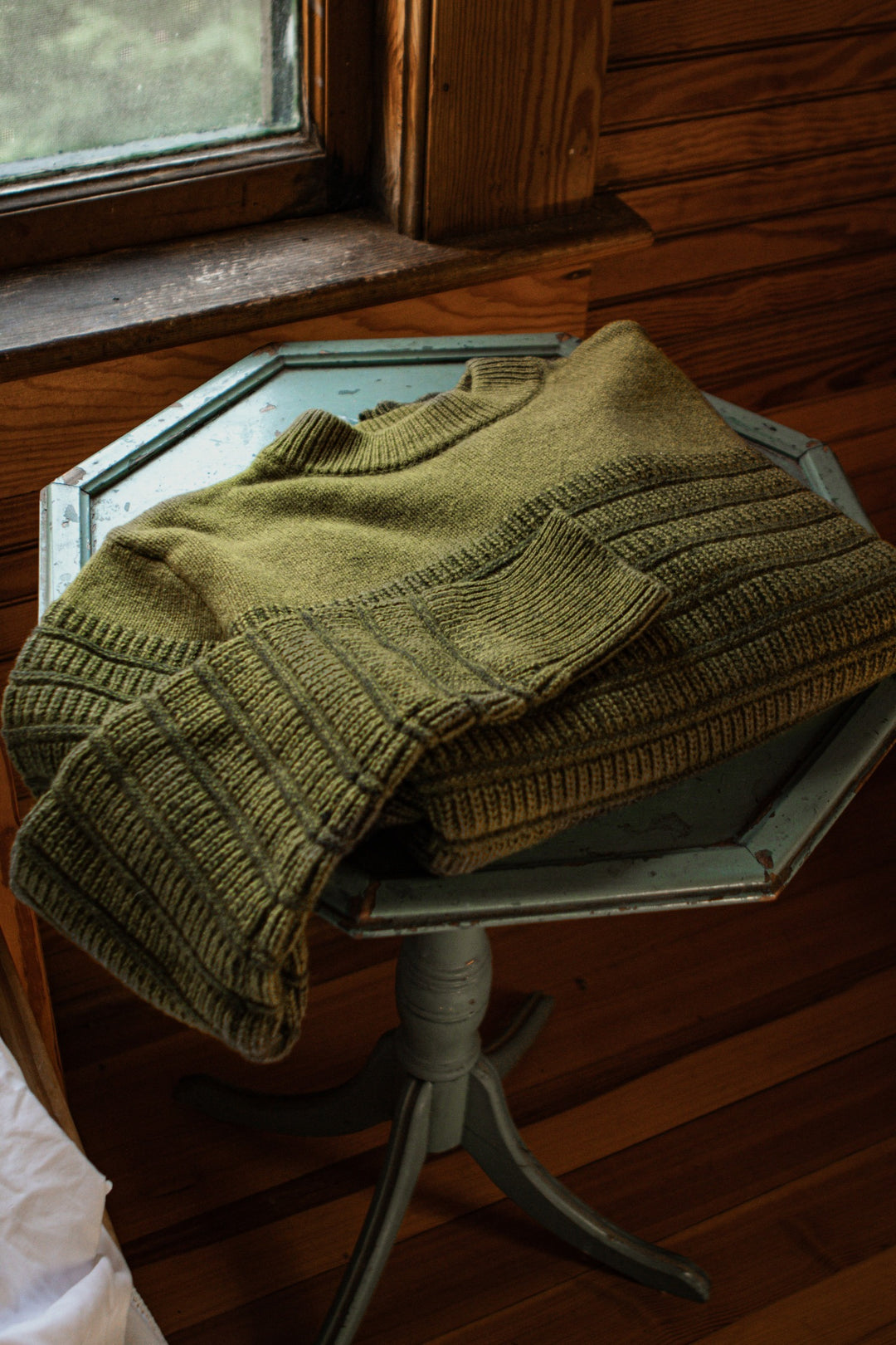 The Bridge & Burn Sabine Sweater in olive sits folded on a small antique wooden table by a window, framed by wood-paneled walls and bathed in warm natural light.