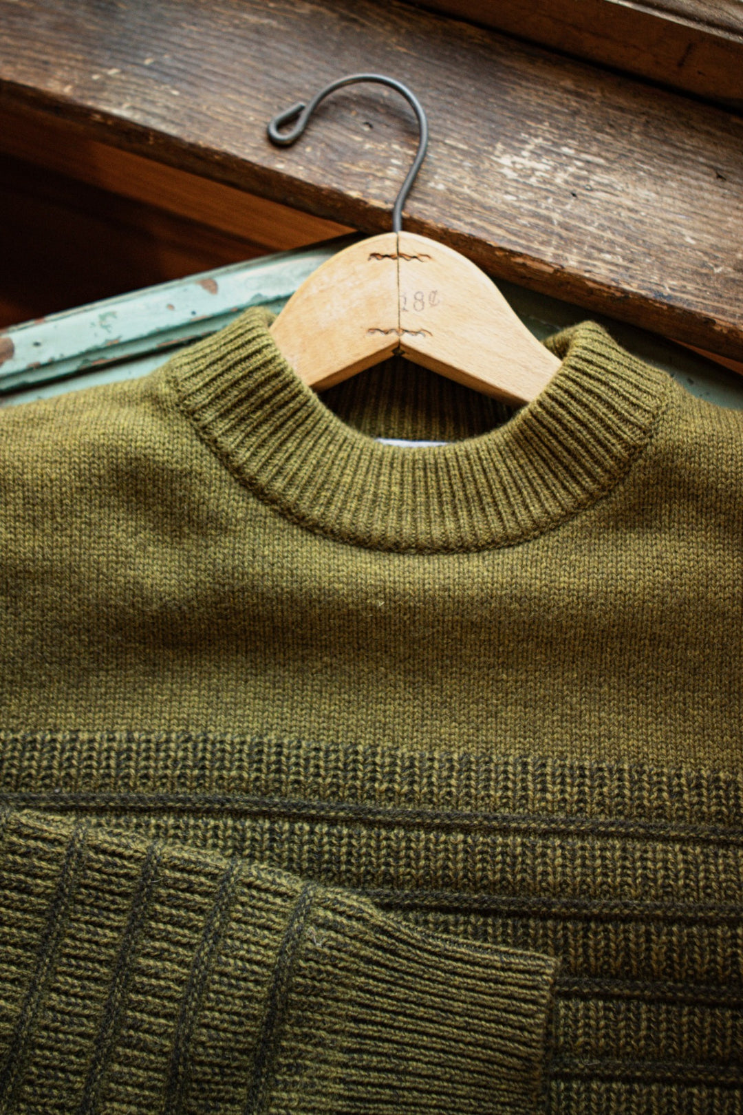 The Sabine Sweater in olive by Bridge & Burn, crafted from merino wool with ribbed cuffs and collar, is neatly folded on a hanger atop a distressed wooden surface—an ideal layering piece for any season.