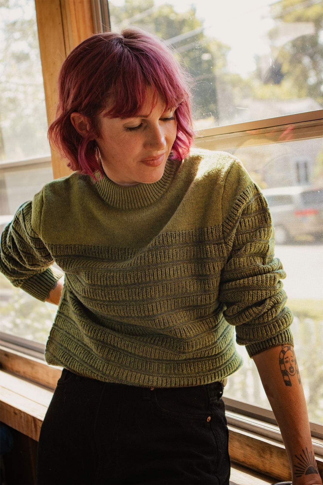 A person with short pink hair and a tattoo on their left forearm stands by a sunny window, thoughtfully looking down while wearing the Bridge & Burn Sabine Sweater in Olive, softly highlighted by natural light.