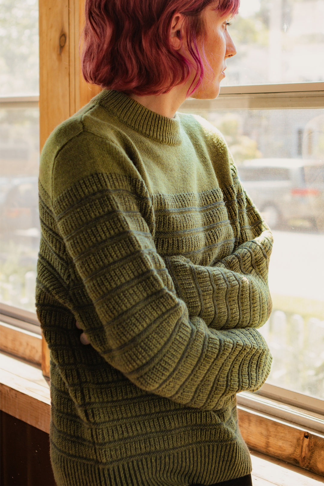 A person with short, wavy, pink hair stands indoors by a sunlit window, arms folded, wearing the Bridge & Burn Sabine Sweater in Olive.