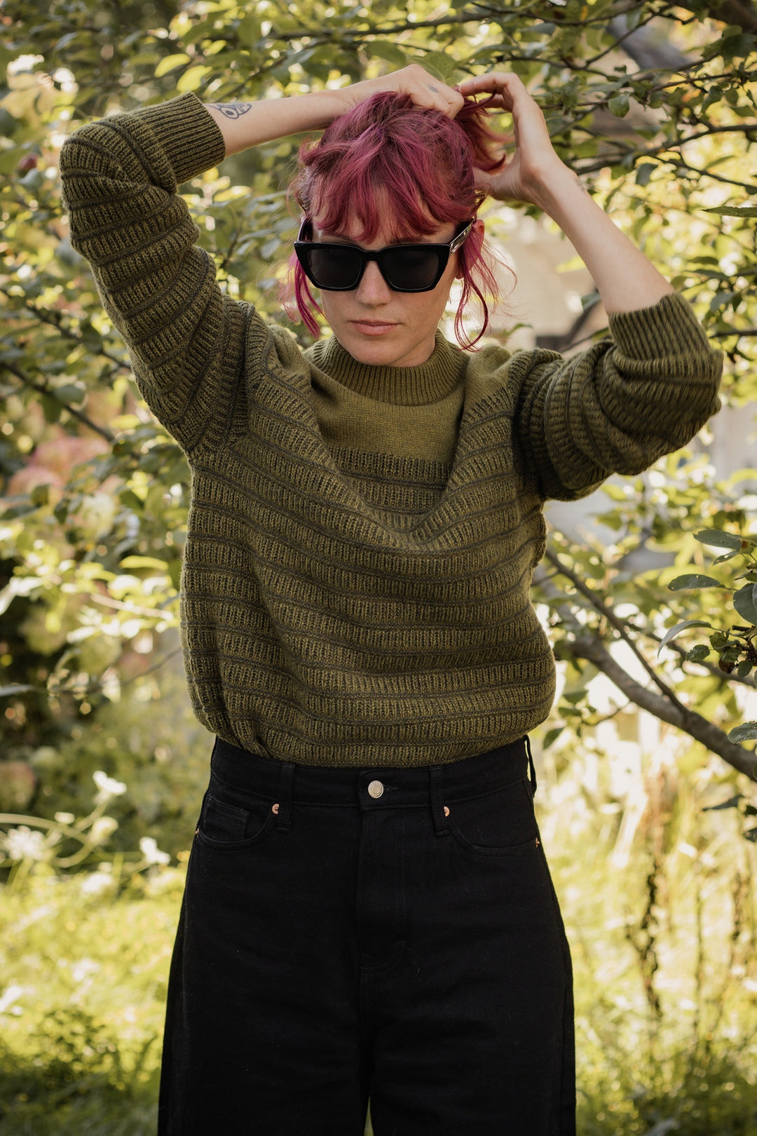 A person with pink hair and sunglasses stands outside, tying their hair. They're wearing the Bridge & Burn Sabine Sweater in Olive with high-waisted black pants, surrounded by leafy greenery in natural light.