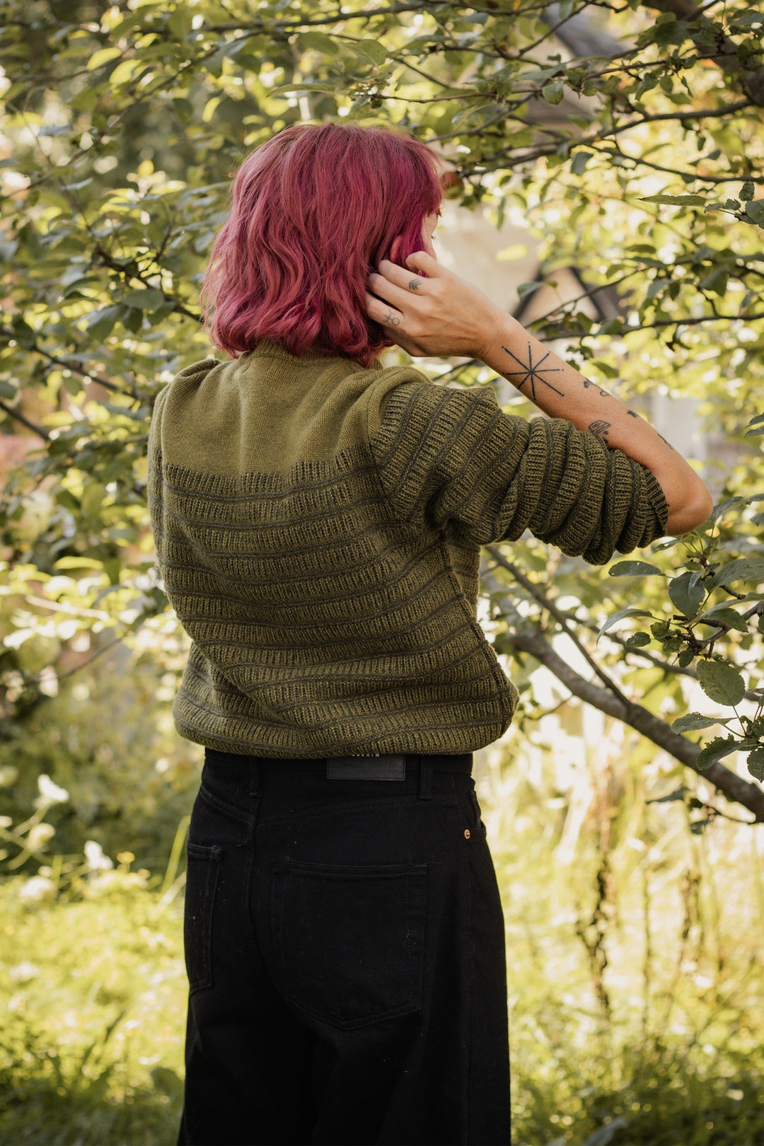 A person with shoulder-length pink hair stands outdoors amid lush greenery, wearing the Bridge & Burn Sabine Sweater in olive and black jeans. One hand touches their head, revealing a geometric star tattoo—perfect for layering.
