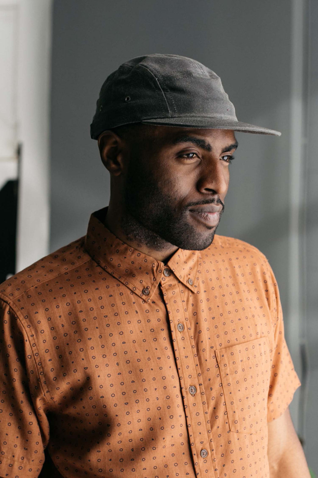 A man wears the Bridge & Burn Camper Waxed 5 Panel Cap in Charcoal and a patterned, short-sleeve orange shirt, standing indoors and gazing thoughtfully to the side in soft, natural light.