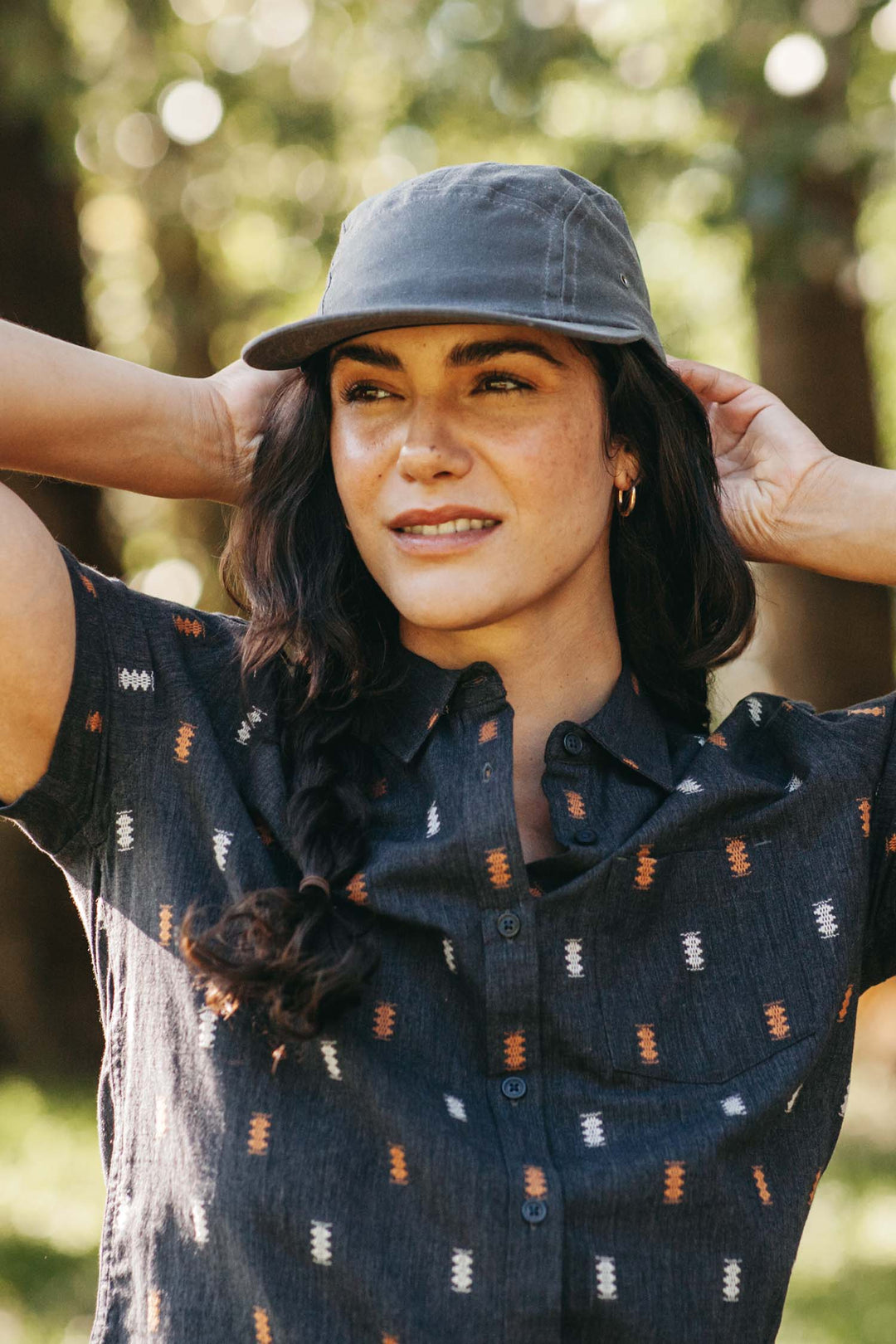 A woman stands outdoors with her hands behind her head, wearing the Bridge & Burn Camper Waxed 5 Panel Cap in Charcoal and a patterned short-sleeve shirt, smiling slightly as sunlight filters through the trees.