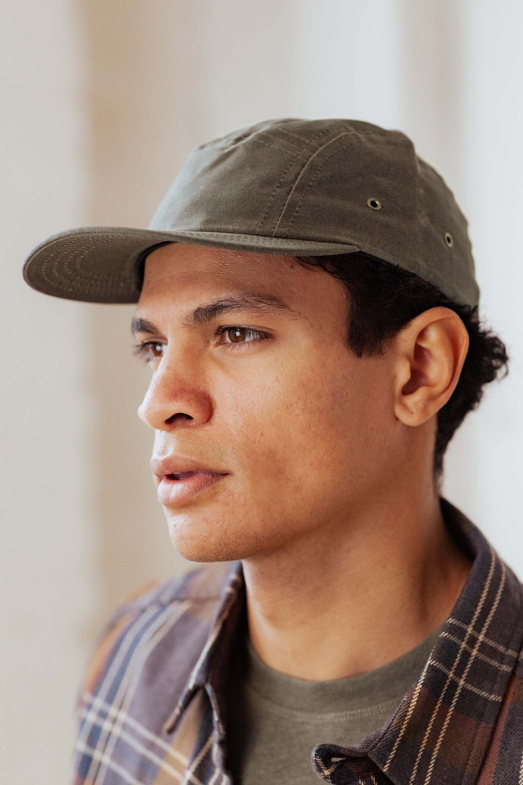A person wearing the Bridge & Burn Camper Waxed 5 Panel Cap in Moss and a plaid shirt looks slightly to the side in soft indoor light.