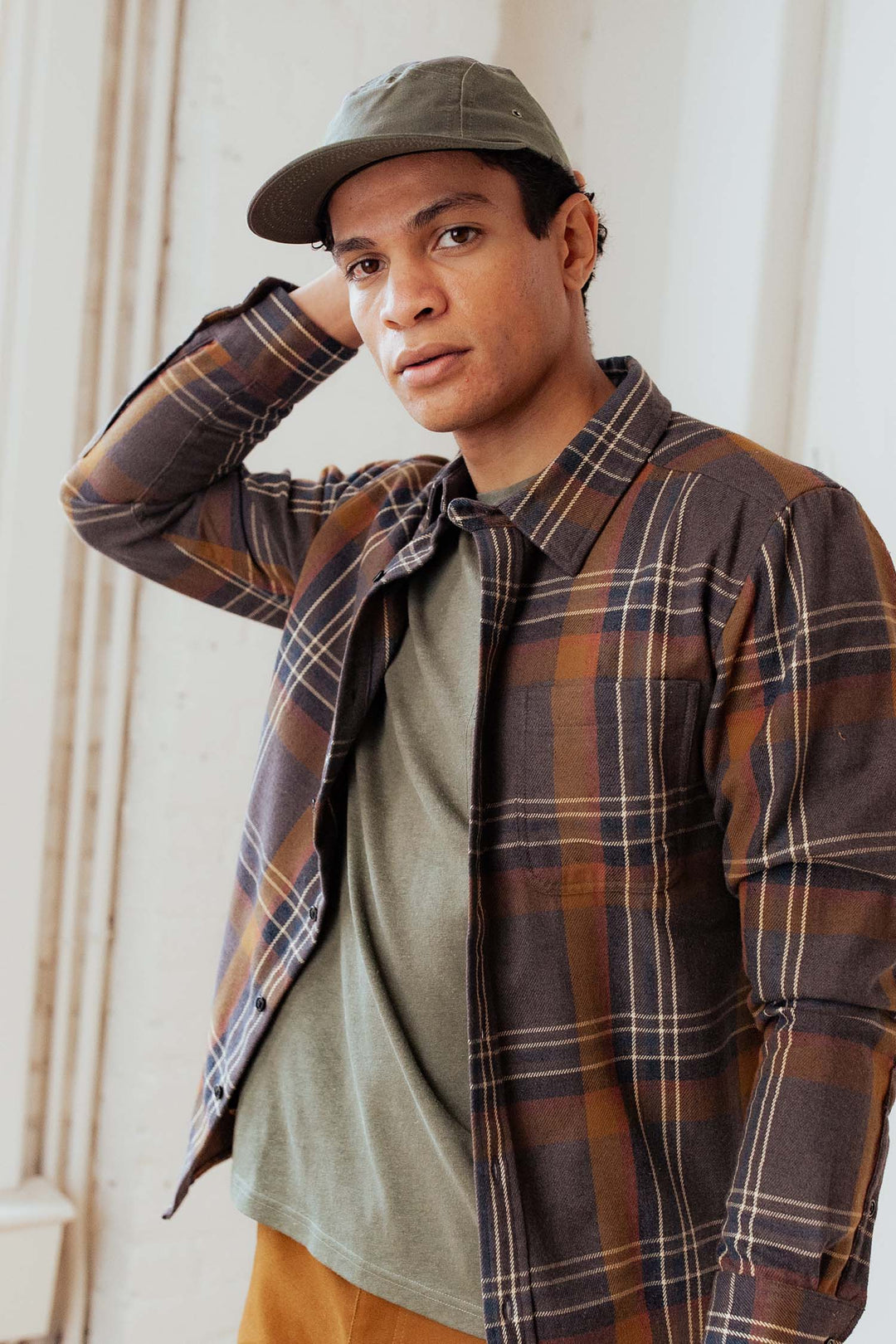 A person wearing the Bridge & Burn Camper Waxed 5 Panel Cap in Moss, an olive green shirt, and a brown plaid jacket stands indoors with one hand behind their head, looking at the camera.