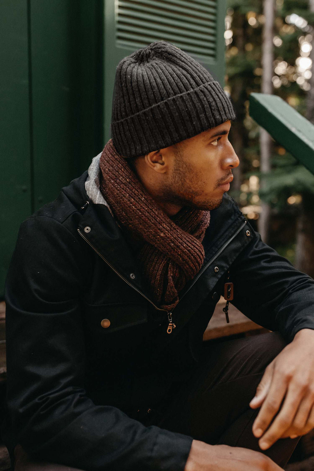 A person in a Bridge & Burn Chunky Ribbed Beanie / Charcoal Donegal, brown scarf, and black jacket sits outdoors against green walls and trees, evoking a cool, forested vibe as they look to the side.