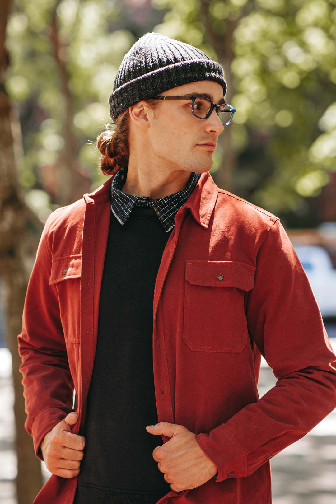 A man in the Bridge & Burn Chunky Ribbed Beanie in Charcoal Donegal stands outdoors, adjusting his jacket and gazing to the side, with trees and warm sunlight softly glowing in the background.