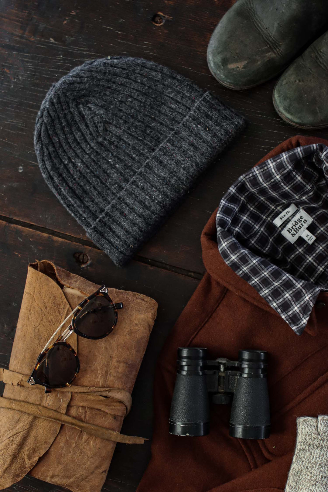 A flat lay of outdoor essentials on a dark wood surface, featuring the Bridge & Burn Chunky Ribbed Beanie in Charcoal Donegal, brown boots, a plaid-lined jacket, black binoculars, round sunglasses, and a rolled leather pouch.