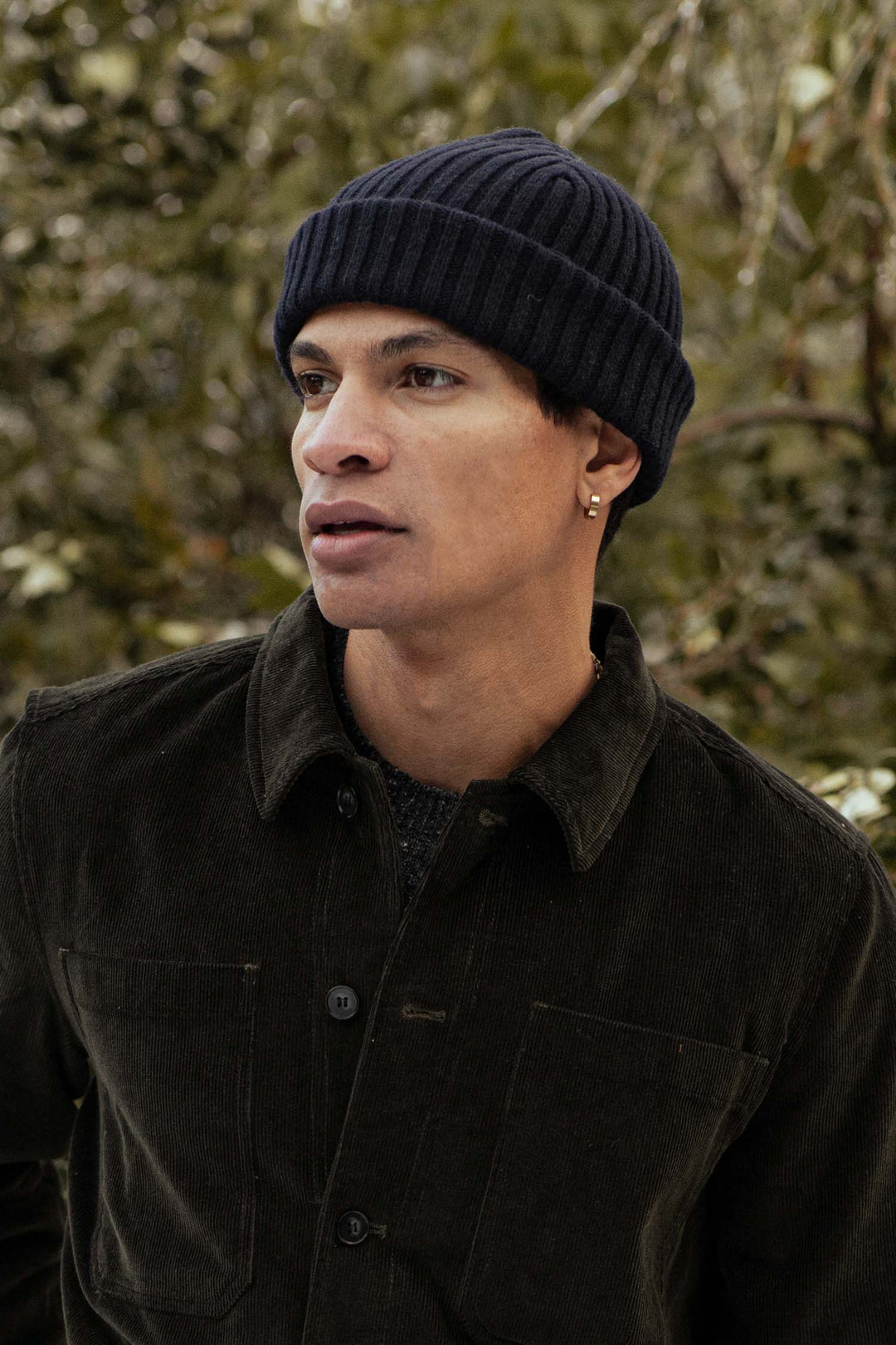 A person wearing the Bridge & Burn Chunky Ribbed Beanie in Dark Navy stands outdoors before blurred greenery, dressed in a dark corduroy jacket and an earring, gazing slightly sideways with a neutral expression.
