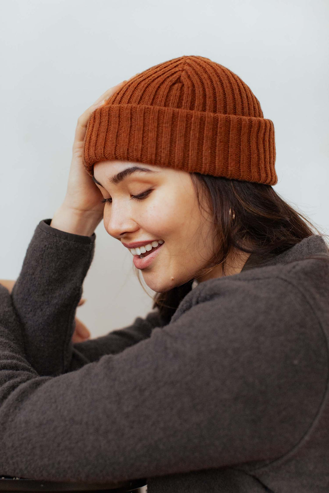 A woman wearing the Bridge & Burn Chunky Ribbed Beanie in Sienna and a dark sweater smiles with her eyes closed, resting her head on her hand against a light background.