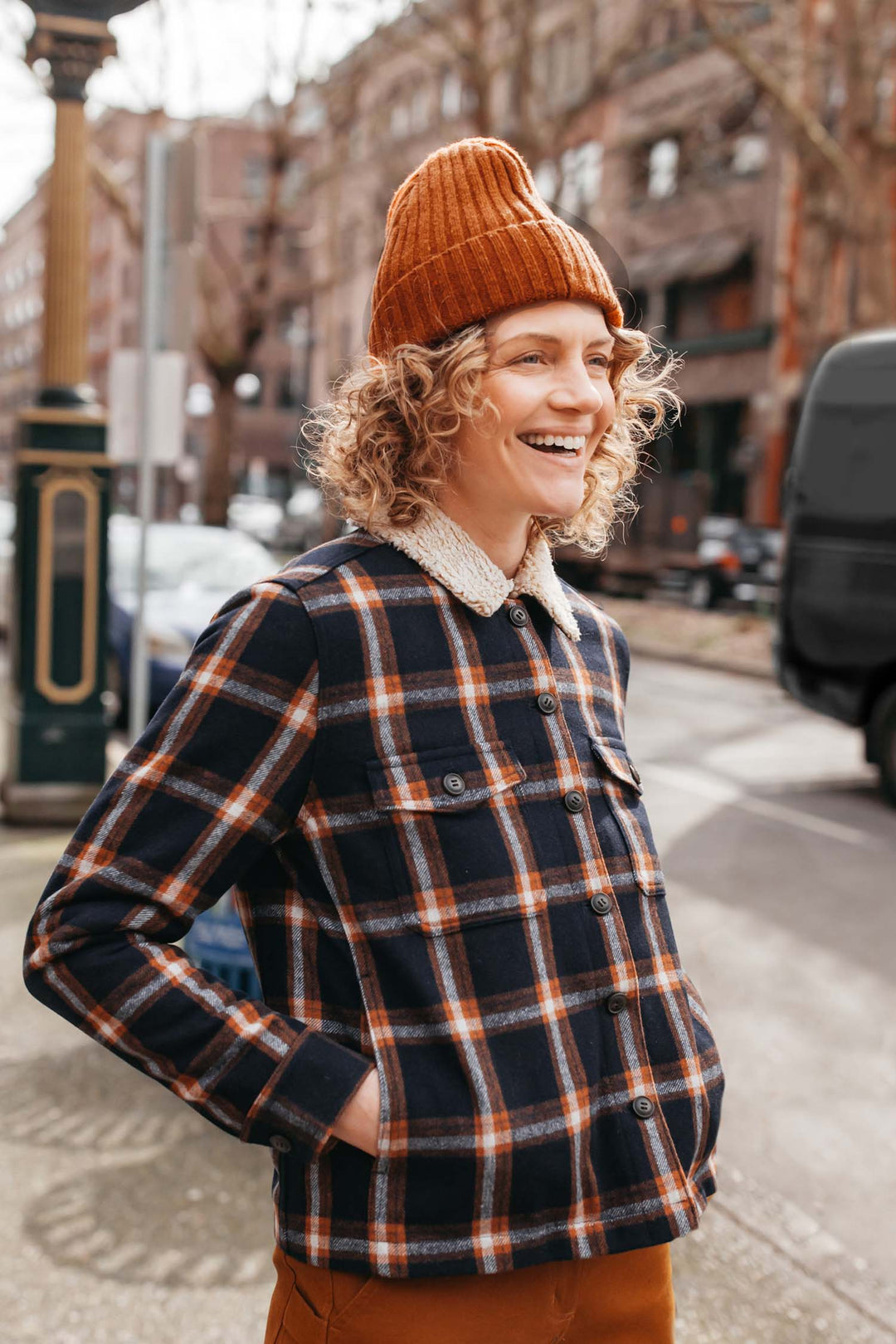 A person with curly hair smiles on a city sidewalk, wearing the Bridge & Burn Chunky Ribbed Beanie in Sienna, a plaid sherpa-collar jacket, and brown pants. Trees and buildings can be seen in the background.